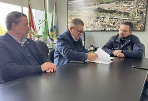 O vice-presidente e diretor de Operações do BRDE, Ranolfo Vieira Júnior, assina contrato em mesa de reunião com o Prefeito de Esteio, Leonardo Pascoal, acompanhados por mais um presente.